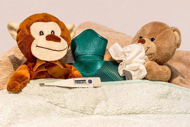 Teddy bears in bed with hot water bottle, thermometer, and tissues, staged to look sick. Travel insurance.