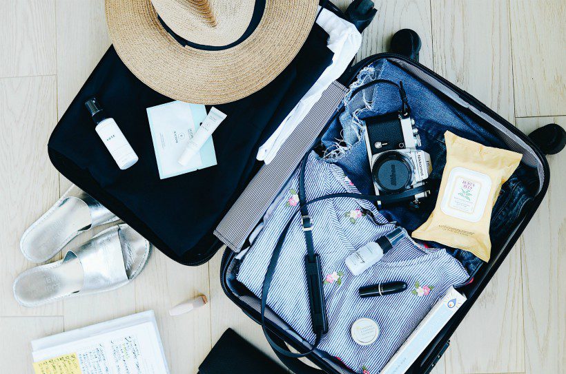 An open suitcase with clothings, camera, hat, and other travel items.