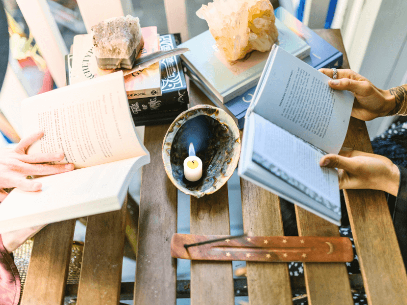 Two pairs of hands holding books, stack of books, a candle, and incense.