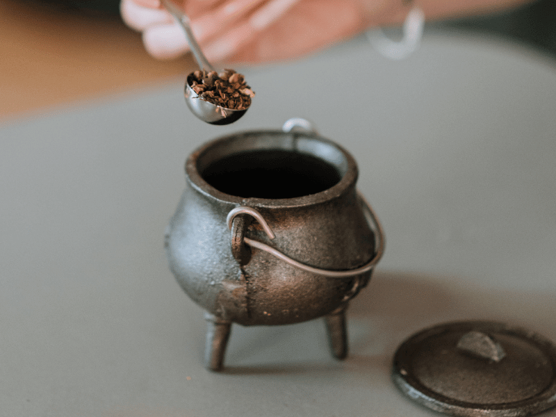 A woman's hand pouring incense resin and herbs into a small cauldron.