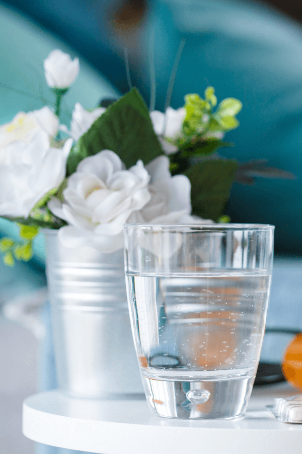 A glass of lowers, flowers, and medication on a bedside table.
