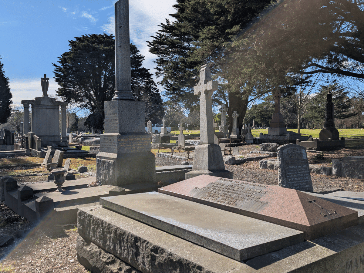 A historic cemetery filled with large stone monuments, crosses, obelisks and family graves beneath mature evergreen trees, with sunlight casting long shadows across the headstones and grass. Fawkner Memorial Park.