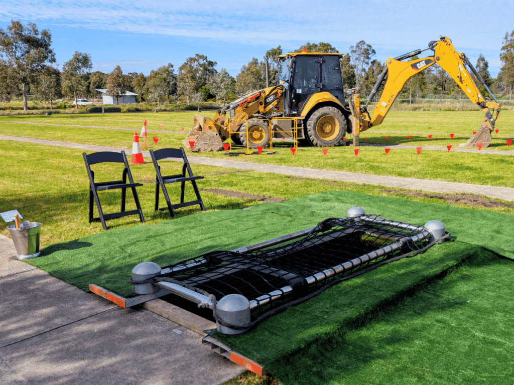 An open grave covered with green artificial turf and a safety net, with two black chairs beside it. A yellow backhoe stands nearby on the cemetery lawn, with the work area marked by orange cones and flags. Fawkner Memorial Park.