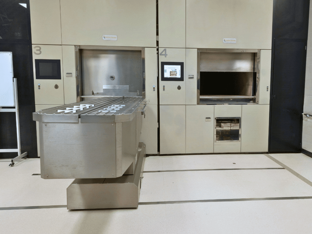 Two numbered cremators built into the wall of a clean, brightly lit crematorium, with one chamber open and a large stainless-steel loading trolley positioned in front.
