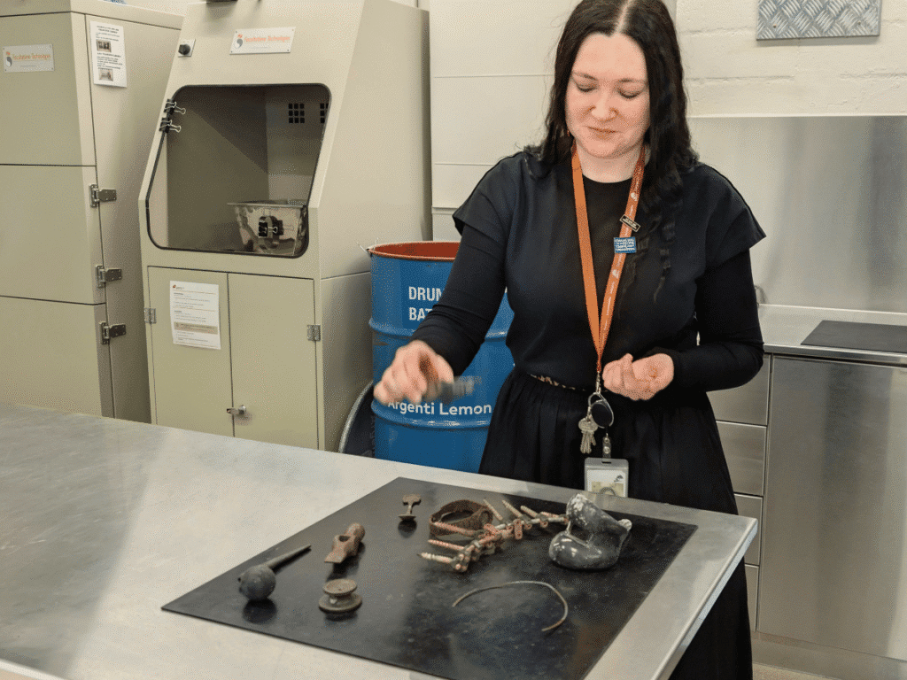 A crematorium operator stands beside a table displaying metal objects recovered after cremation. A cremulator is visible behind her.