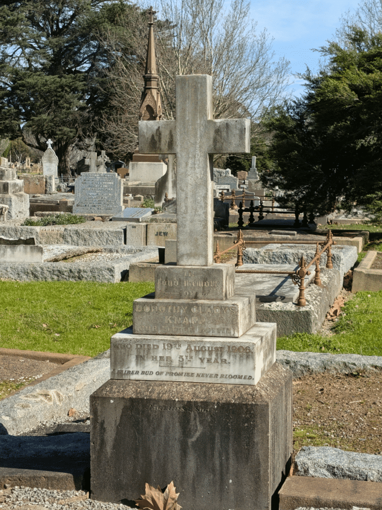 An old cemetery grave marked by a large weathered stone cross, surrounded by historic headstones, iron grave railings and mature trees. The inscription commemorates Dorothy Gladys Knapp, who died in 1906 aged five. Fawkner Memorial Park.