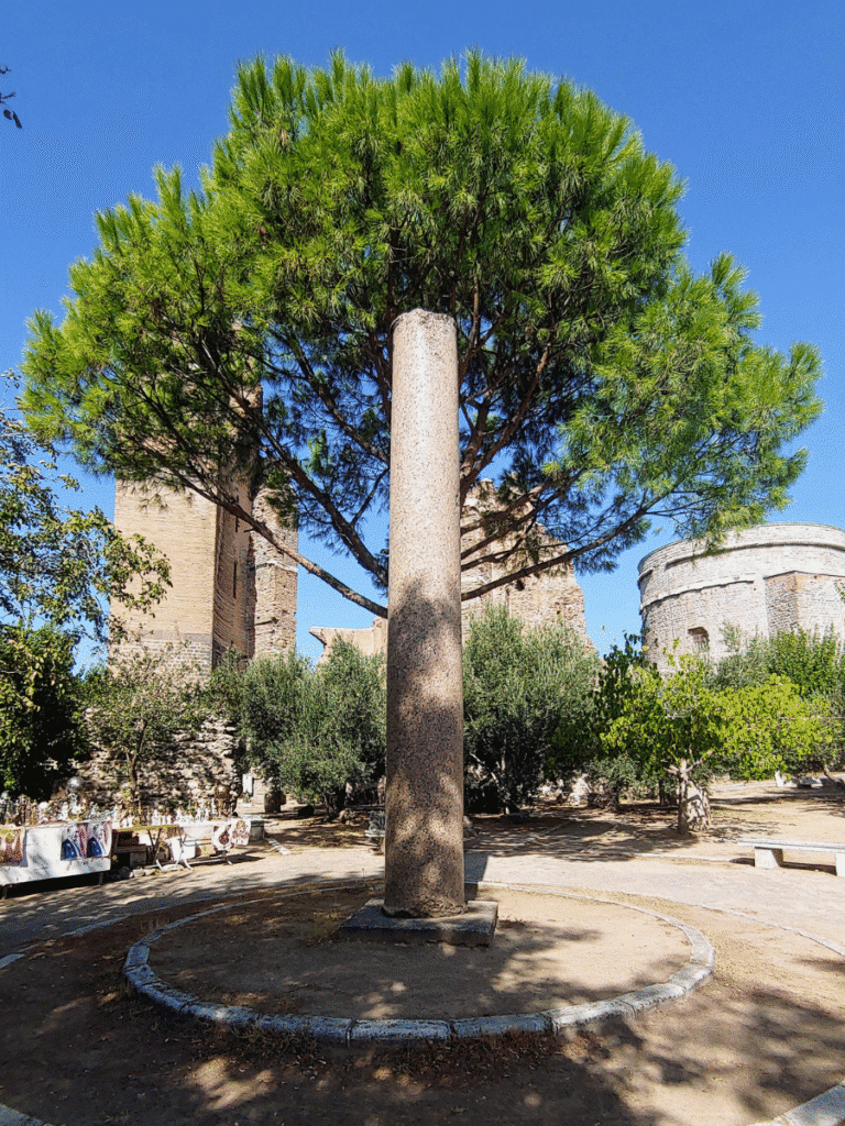 Tall monolithic stone column beneath a large pine tree, with the Red Hall ruins and a rotunda behind it.